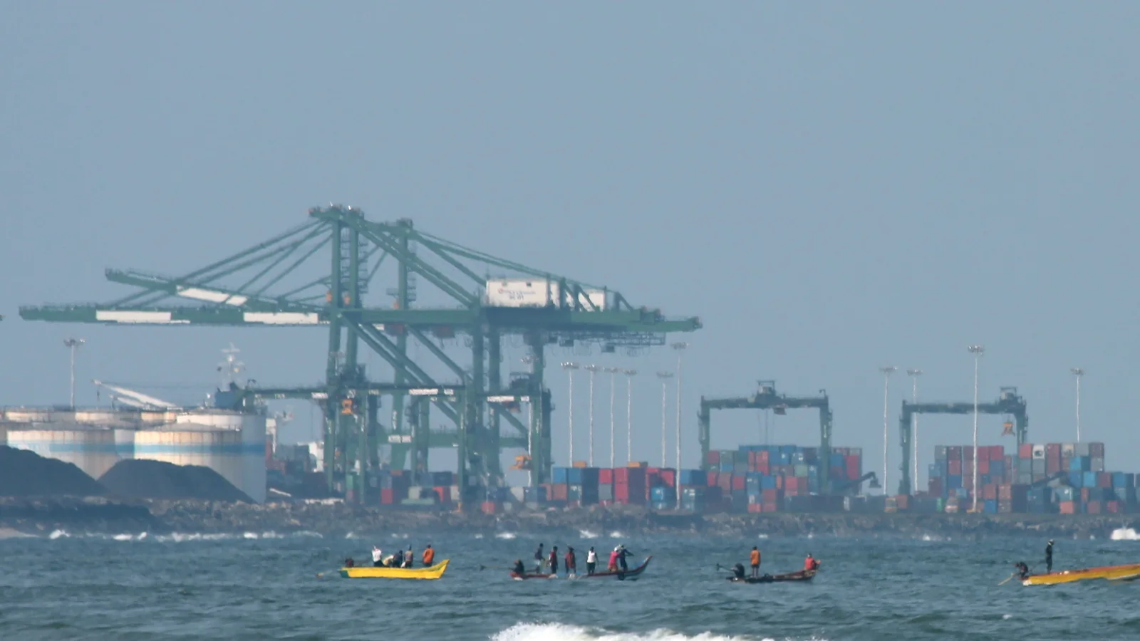 Container cranes and stacked boxes at Chennai Port, seen across the water from Marina Beach, with fishing boats in front.
